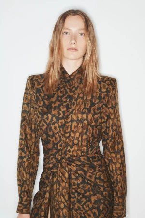 A woman in a long-sleeve animal print dress poses against a white background in a studio setting. The dress is brown and black.