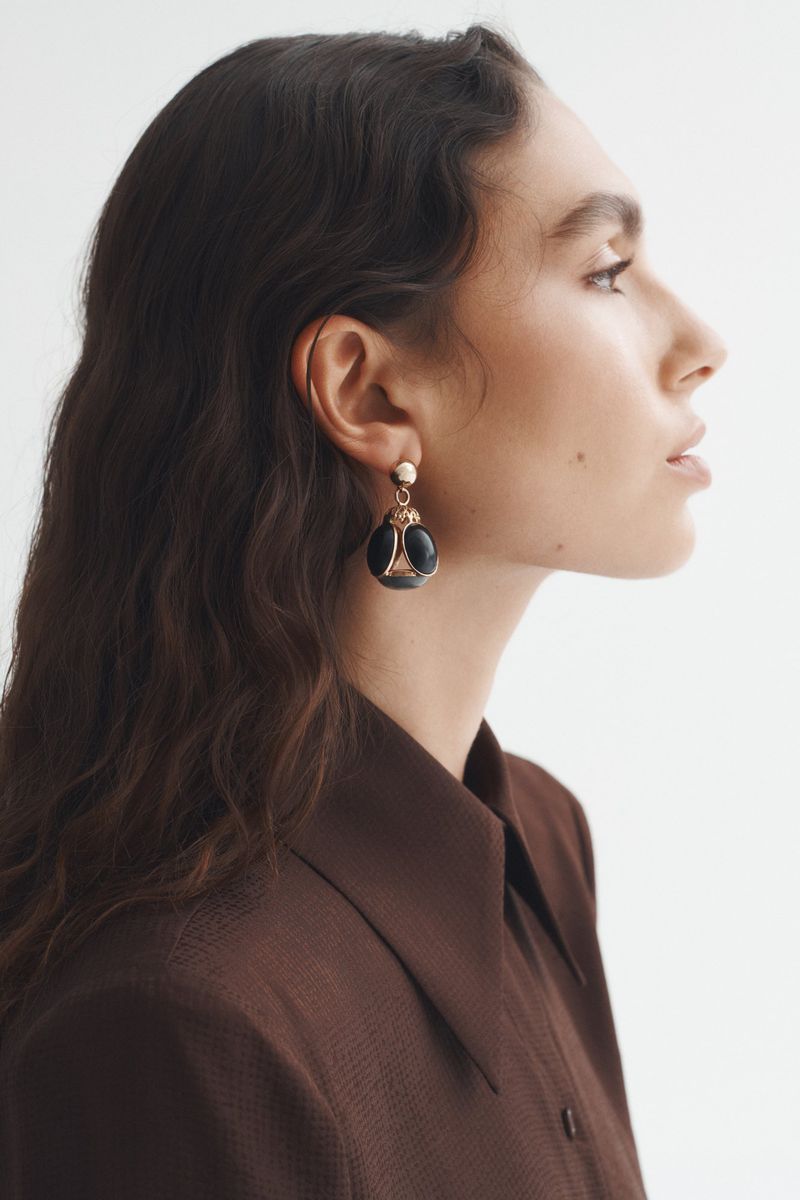 Profile portrait of a woman with dark wavy hair wearing black and gold earrings and a brown collared shirt against a white background.