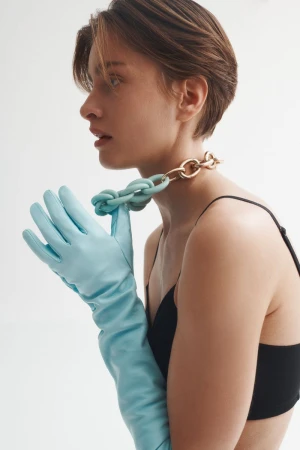 A woman poses in a studio with a light skin tone, short brown hair, a necklace, black top, and long light blue gloves, eye-level shot.
