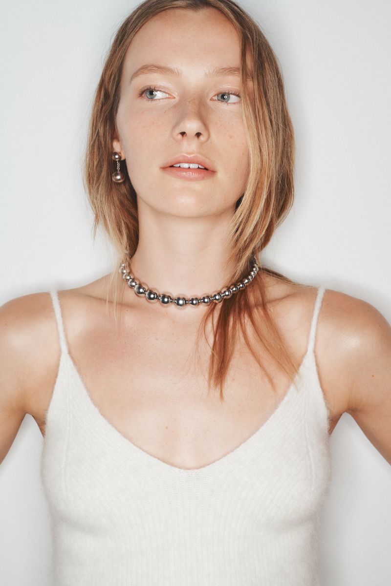 Young adult woman in a white knit tank top wearing a silver beaded choker and matching drop earrings against a white background.