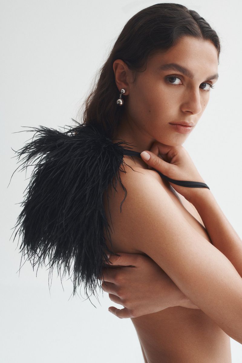 Portrait of a woman with dark hair wearing a black feathered shawl, posing against a clean white backdrop in a beauty editorial.