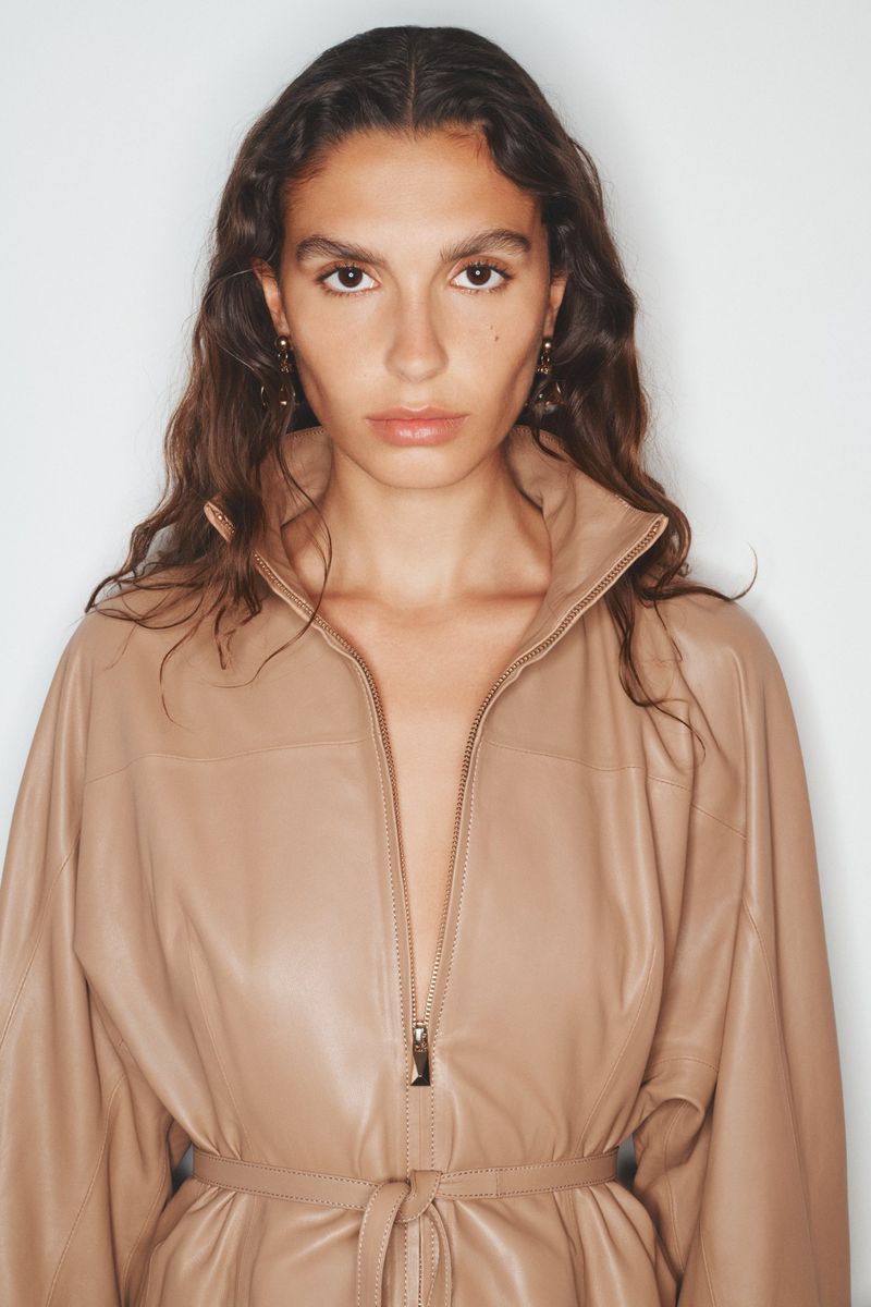 Close-up shot of a tan-skinned model in a beige leather jumpsuit. She poses in a photo studio with soft lighting and long wavy brown hair.