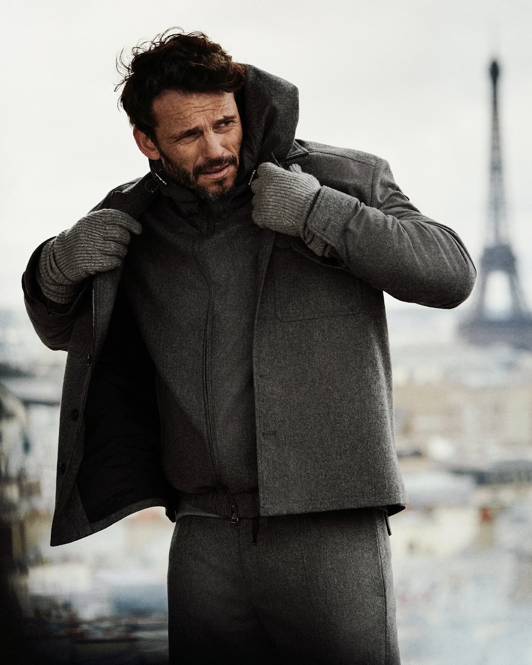 Man in grey wool jacket, zip-up sweater, gloves, and trousers poses outdoors with Eiffel Tower in the distance on a cloudy day.