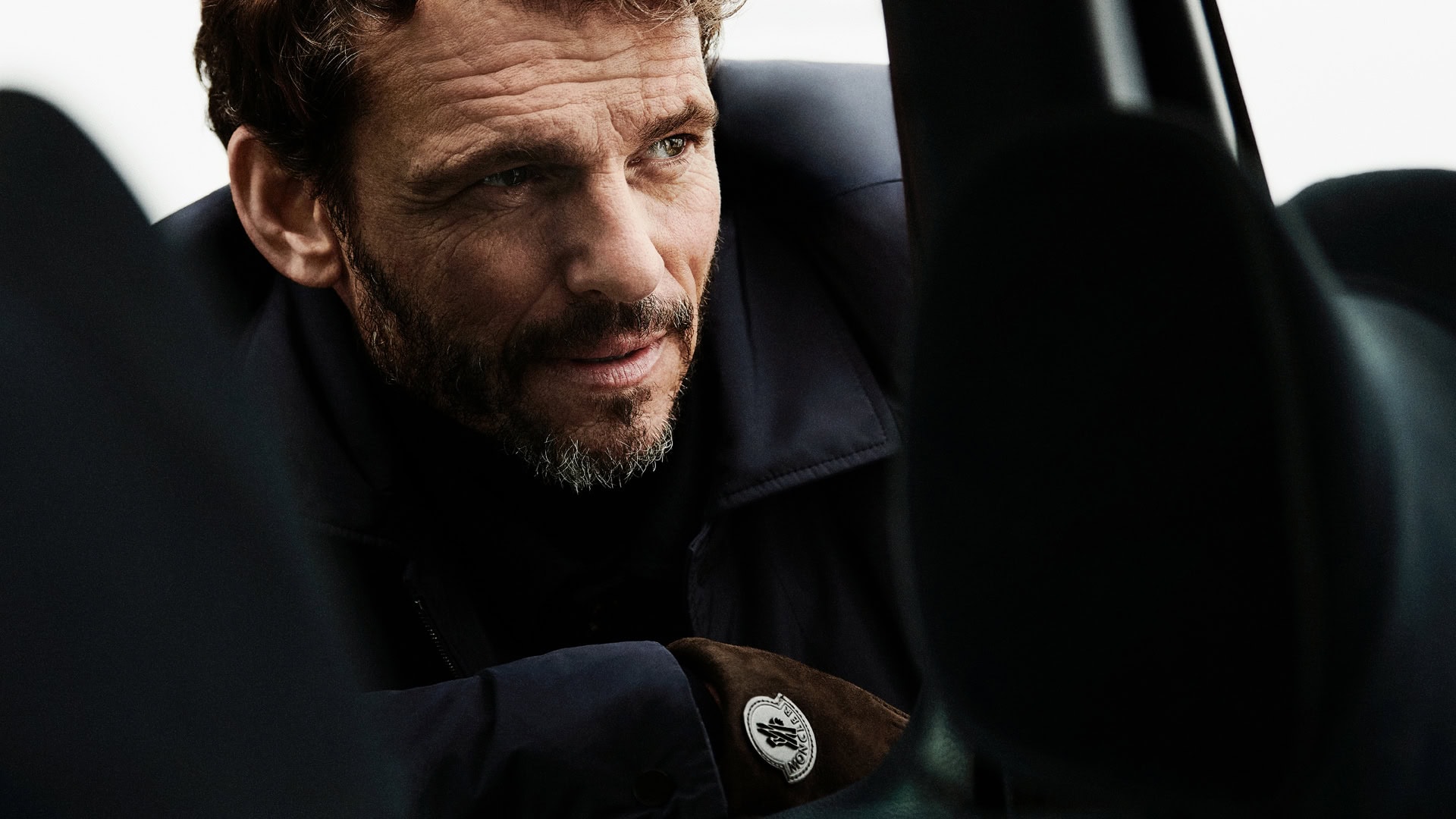 Mature man in a navy coat with a logo patch on his suede glove, inside a car with black leather upholstery, looks away.