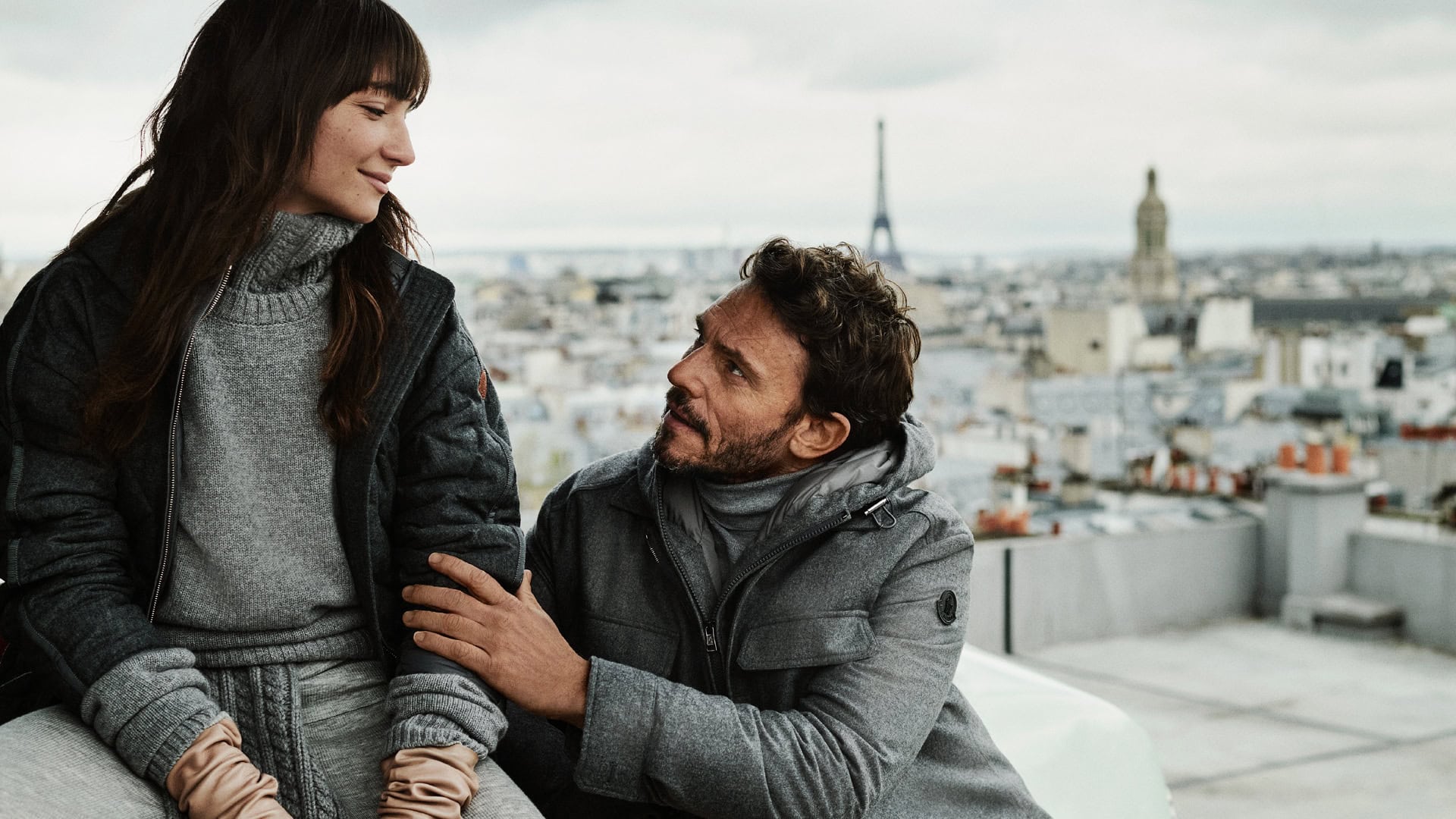 A couple poses on a rooftop in Paris, France. The man gazes at the woman, who looks into the distance. The Eiffel Tower is visible.