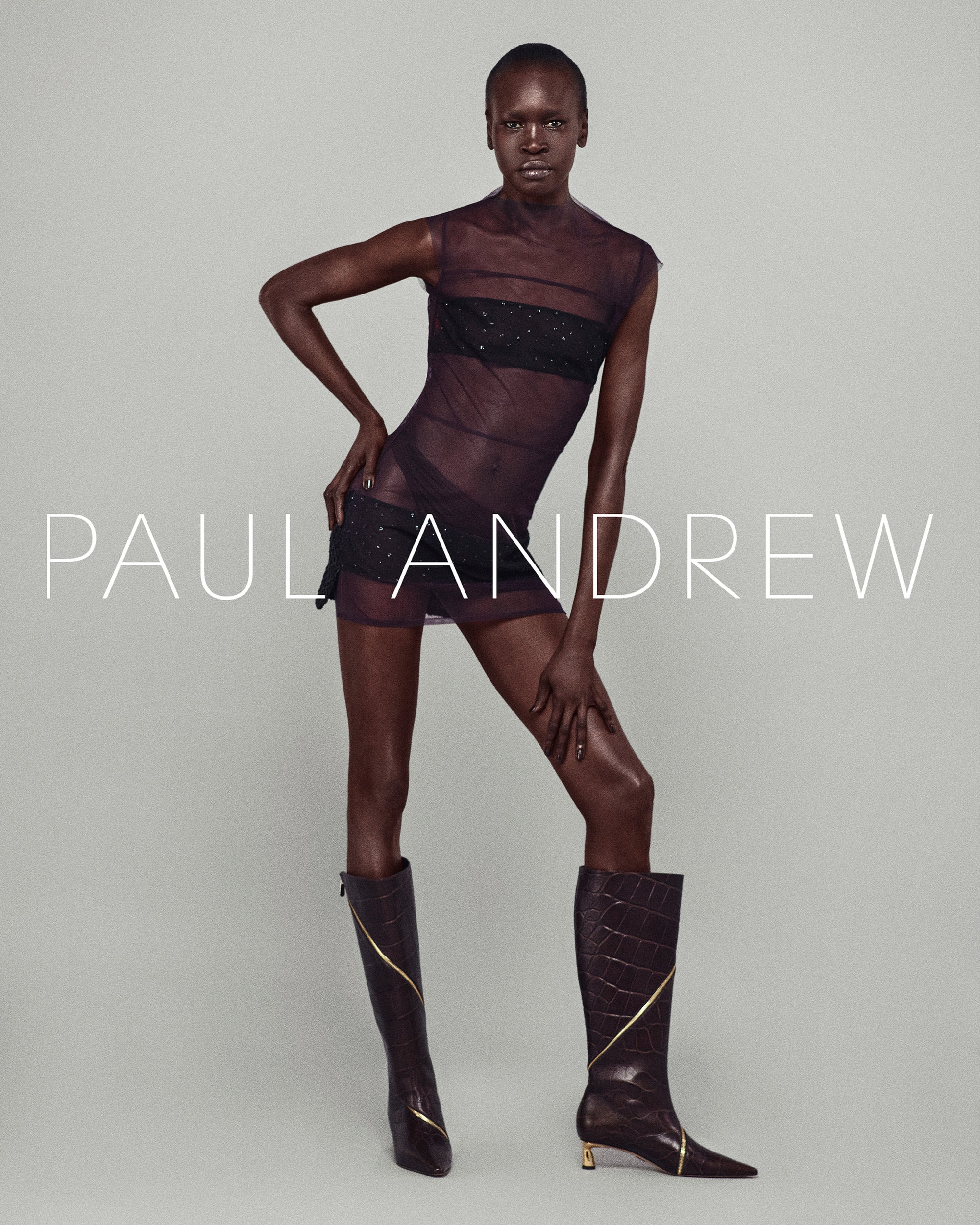 Model Alek Wek in a sheer dark dress and tall boots against a neutral backdrop for Paul Andrew