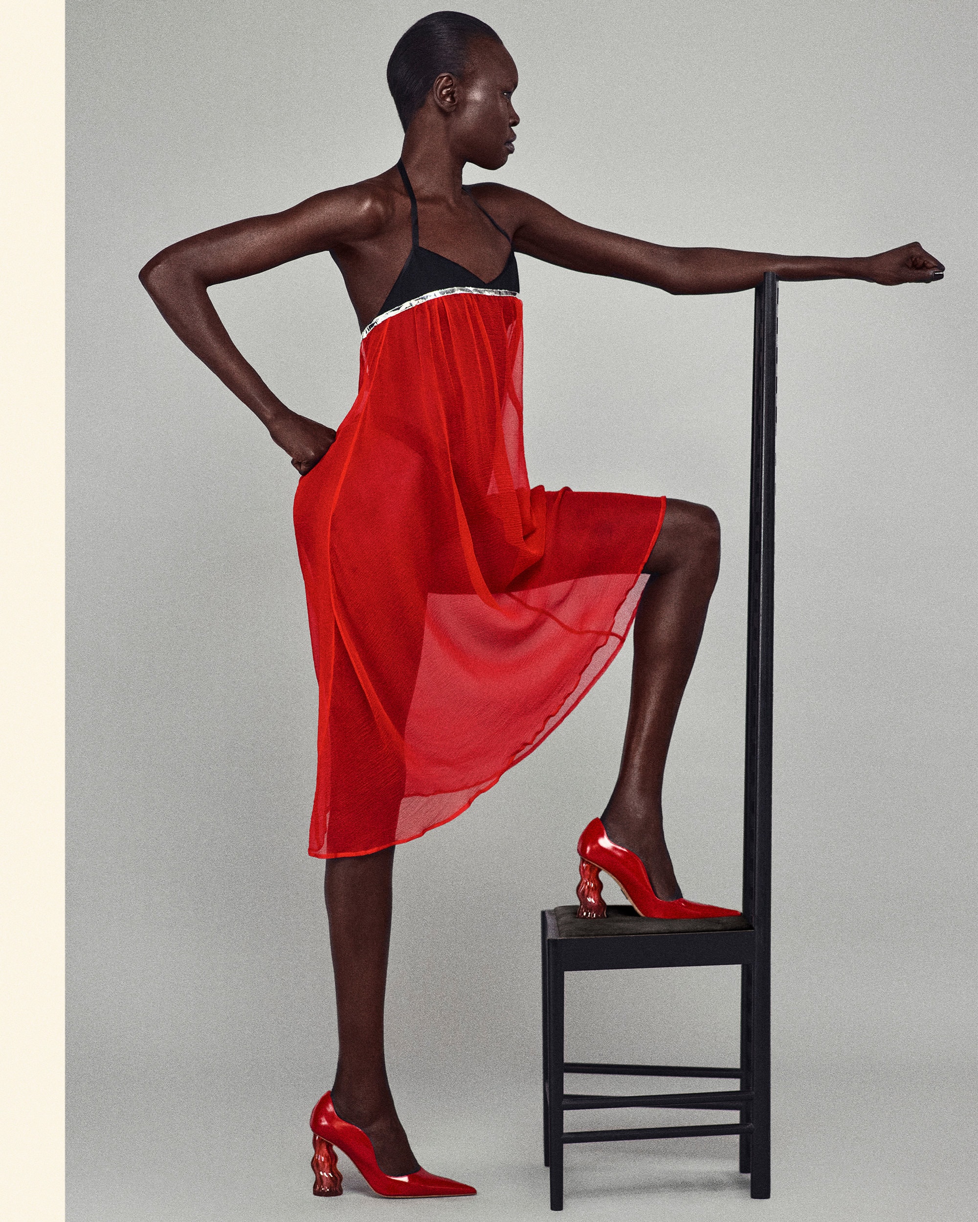 Fashion shot of a Black female model in a red chiffon dress and red heels, posing next to a black chair in a studio setting.
