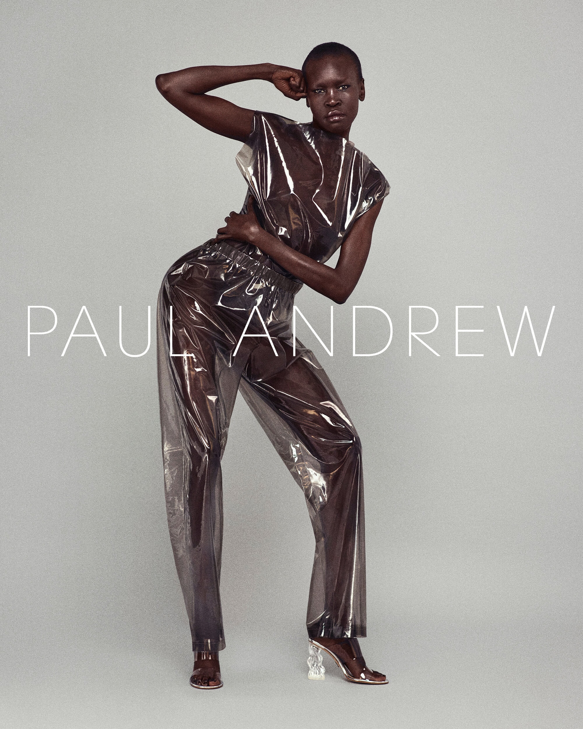 Fashion shot of an adult female model wearing transparent plastic pants and top with high heels, posing in front of a grey background.