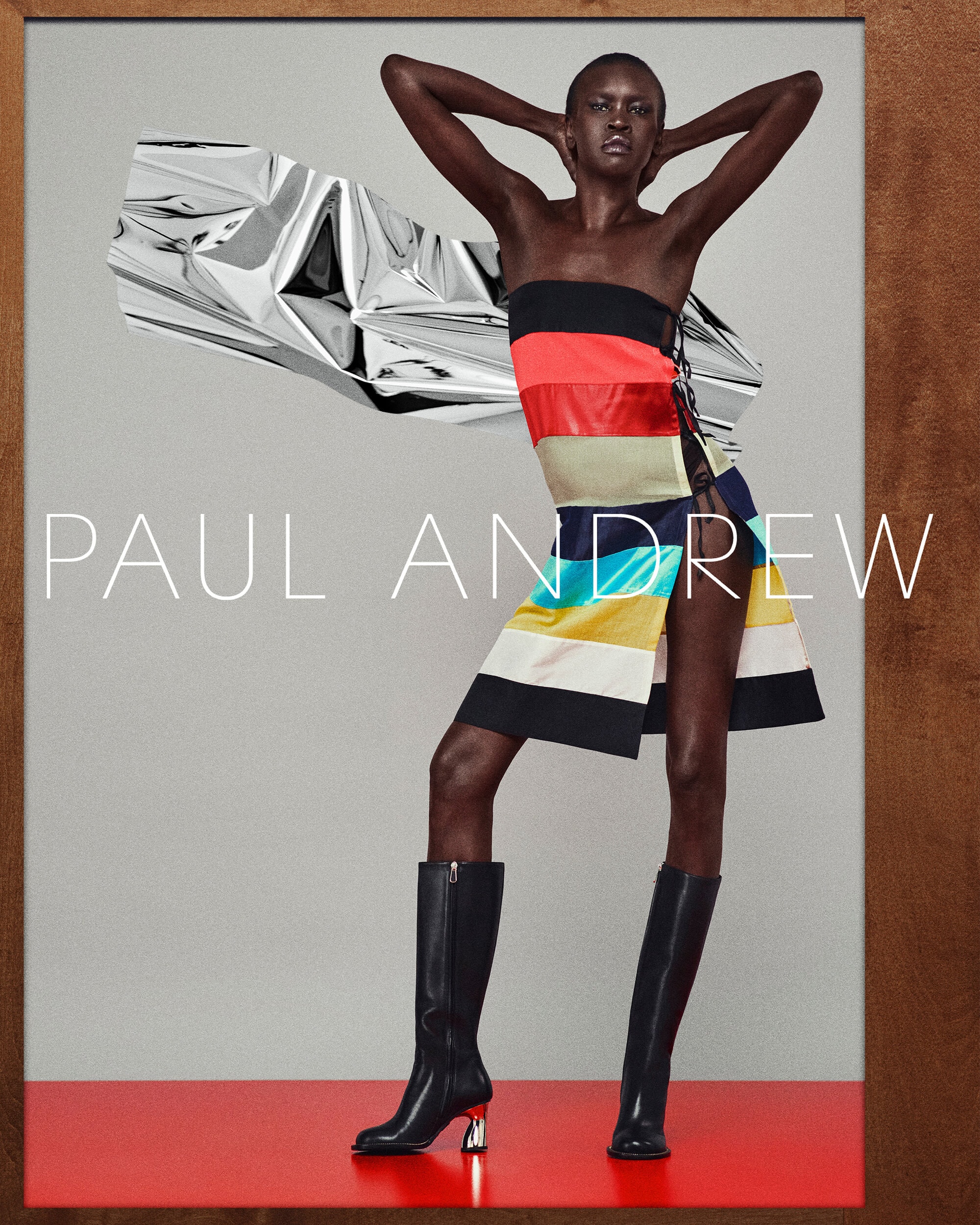 Fashion shot of a slim, dark-skinned woman posing in a colorful dress and boots against a studio backdrop with silver fabric art piece.