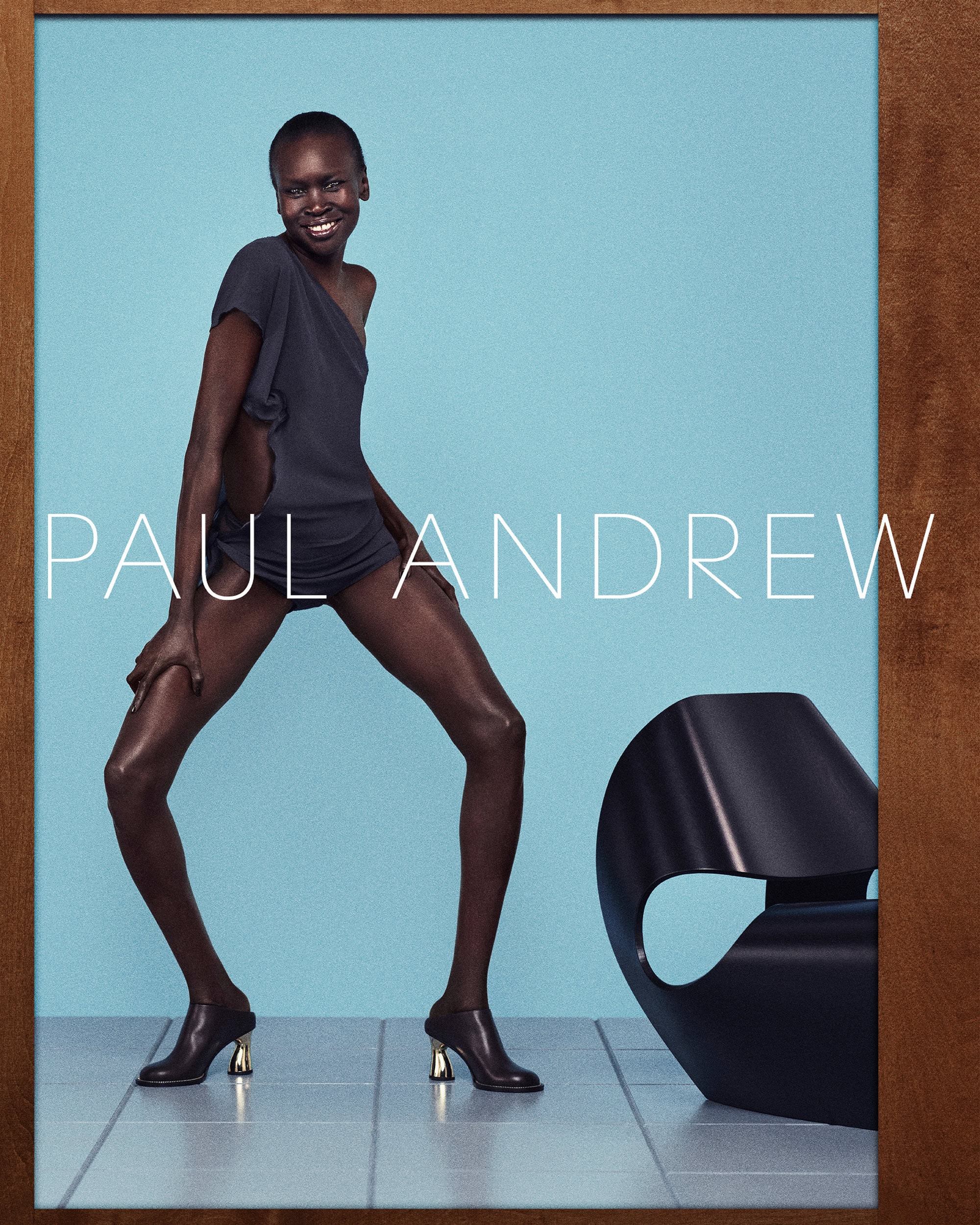 Alek Wek wearing dark top and heeled mules posed against a bright blue studio backdrop for Paul Andrew campaign.