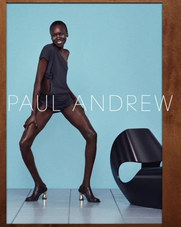 Alek Wek wearing dark top and heeled mules posed against a bright blue studio backdrop for Paul Andrew campaign.