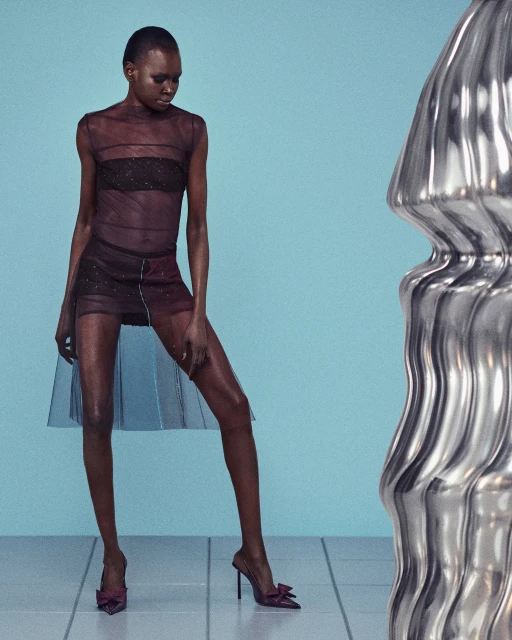 Elegant adult female in sheer dress and heels posing in a photo studio with a silver sculpture against a light blue backdrop.