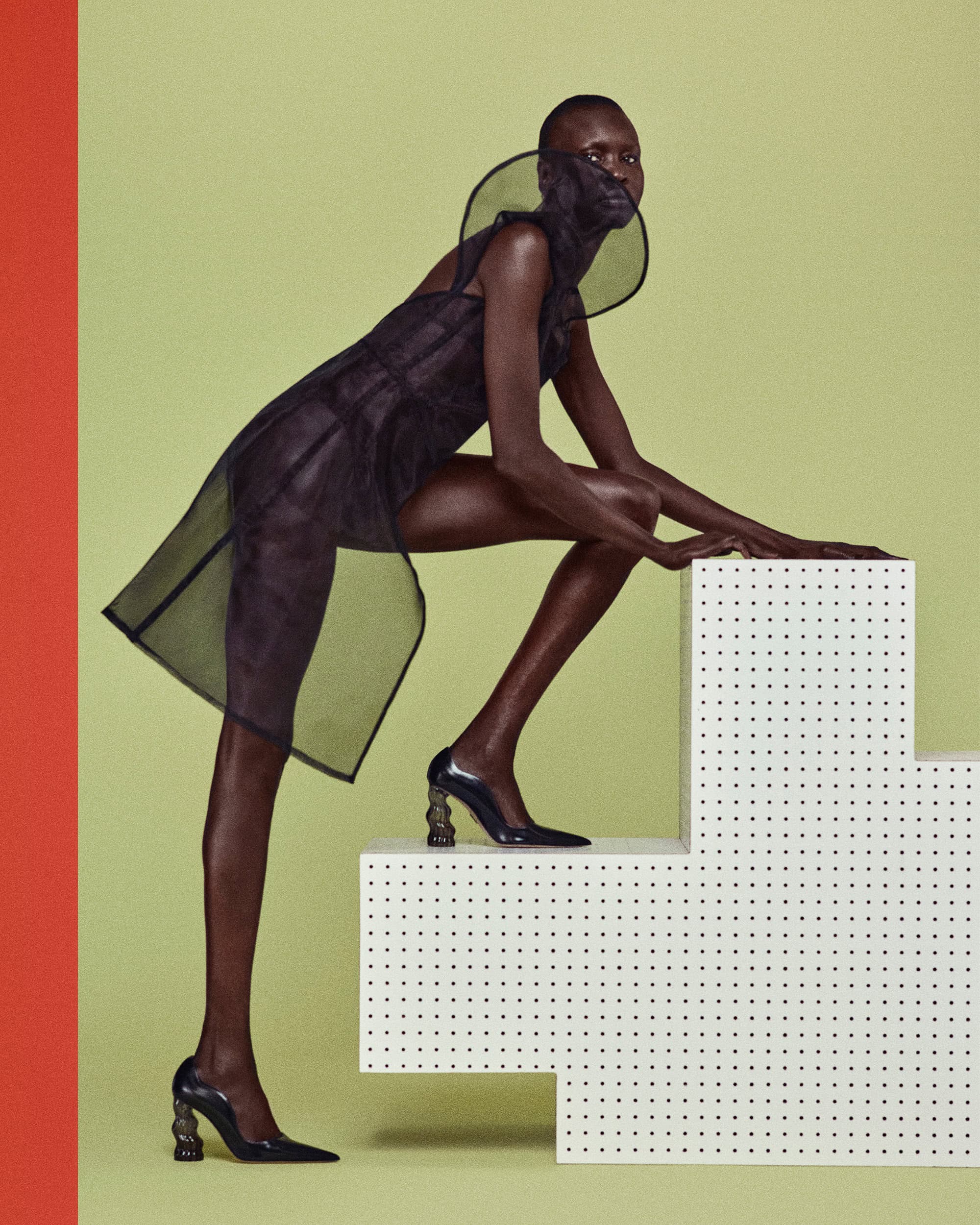 Fashion photo of a Black female model in a translucent black dress and sculptural heels, posing on white stairs against a light green backdrop.