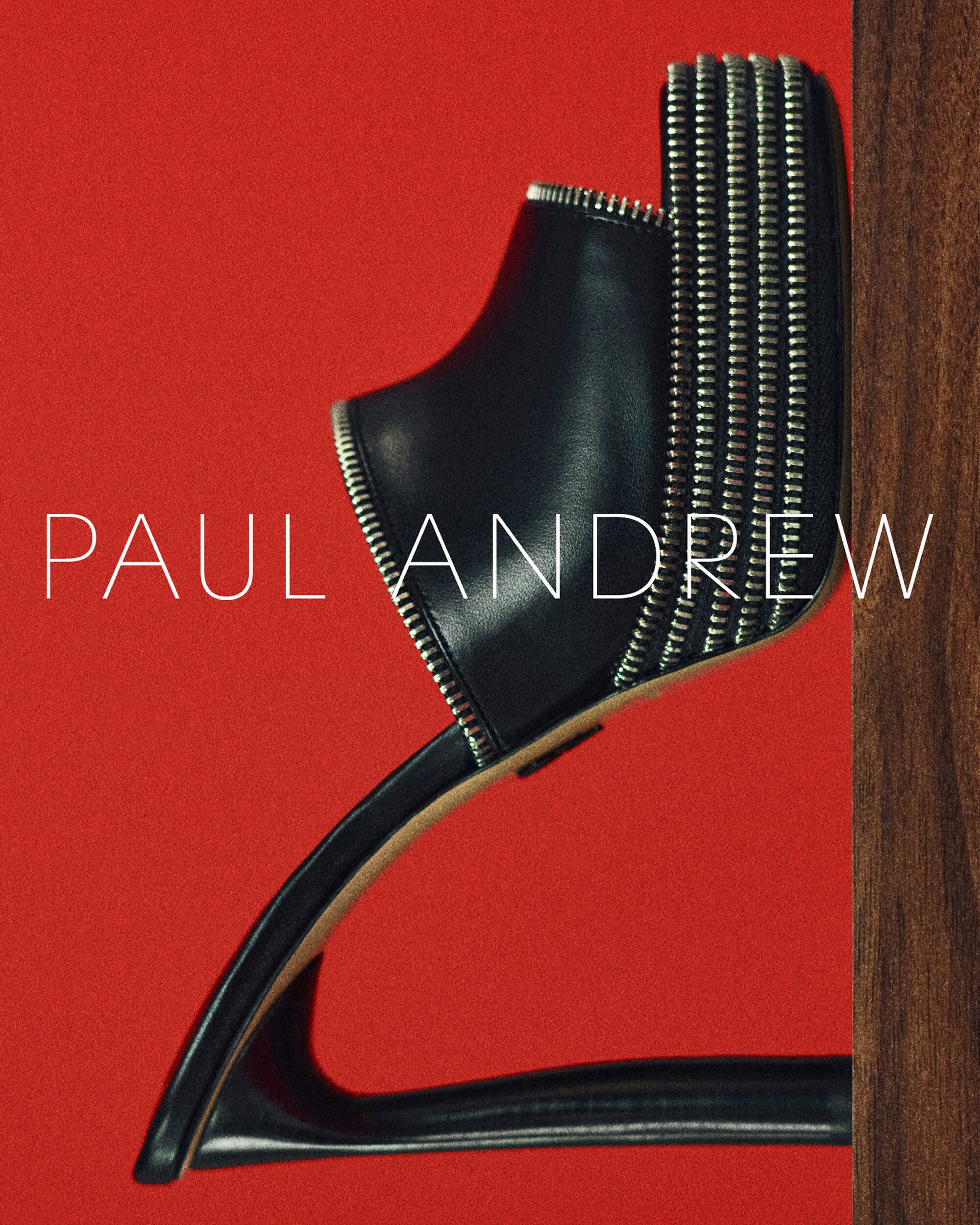 Studio shot of a single black leather heeled mule shoe with zipper detailing against a red background with a dark wood element.