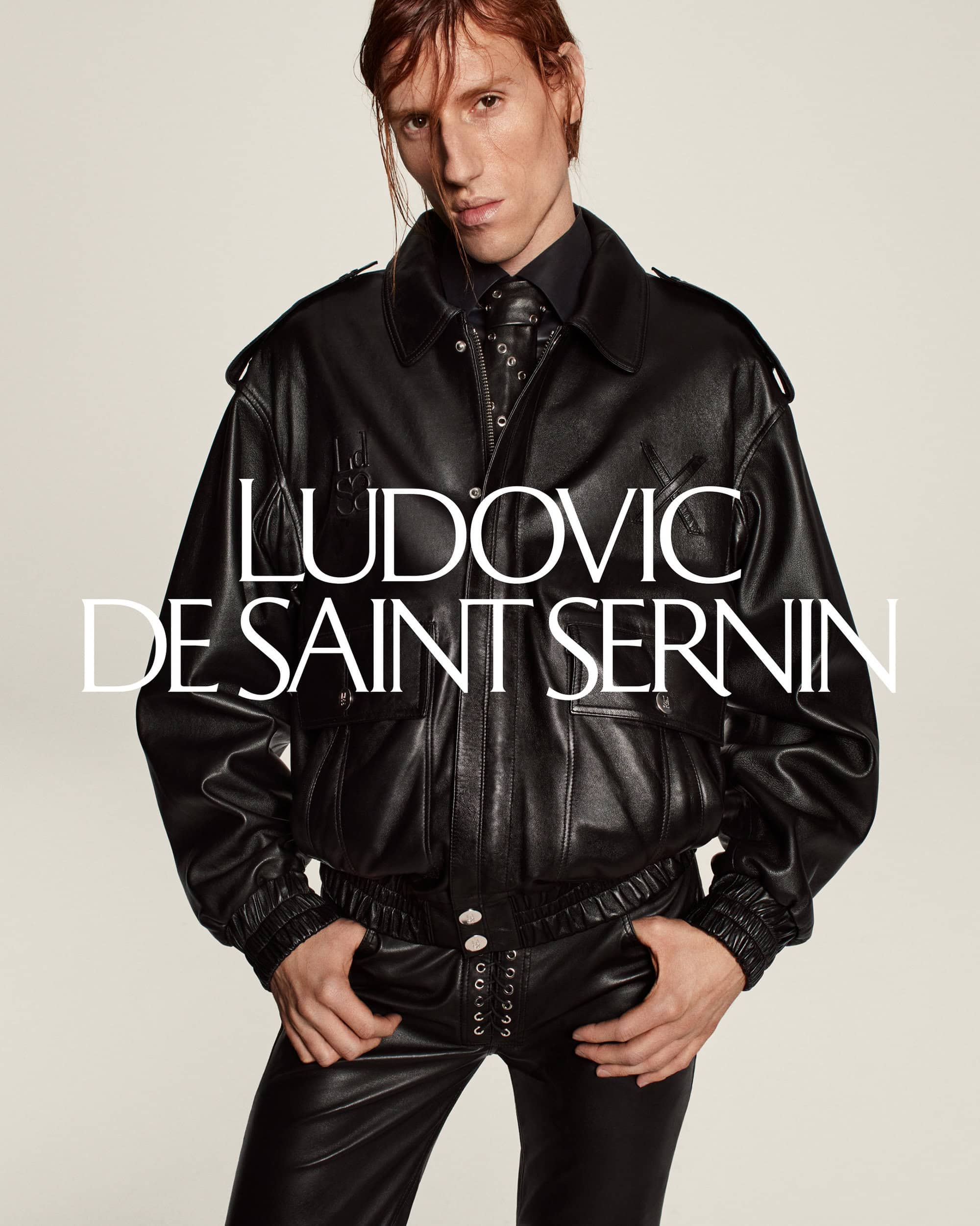 Fashion shot of a slender male model wearing a black leather jacket and pants, standing against a clean background with brand logo.