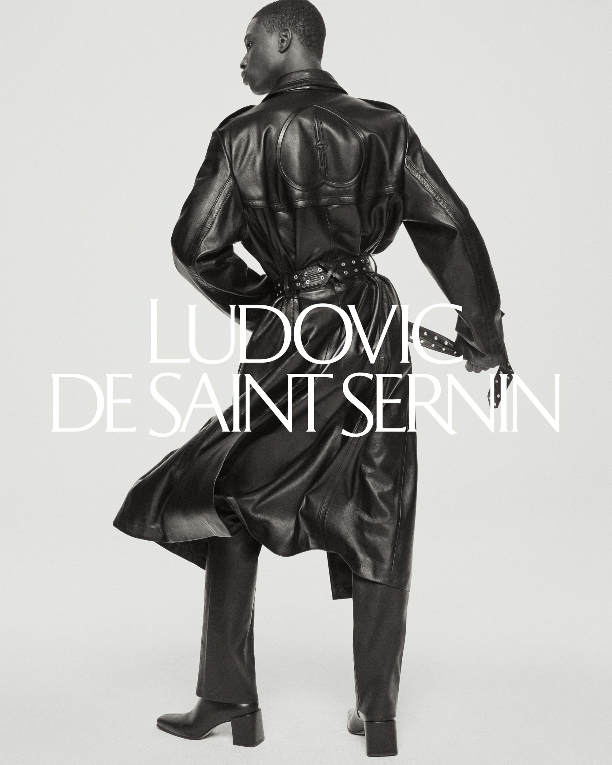 Model showcasing a black leather trench coat, trousers, and boots by Ludovic de Saint Sernin, posing in a studio against a plain background.