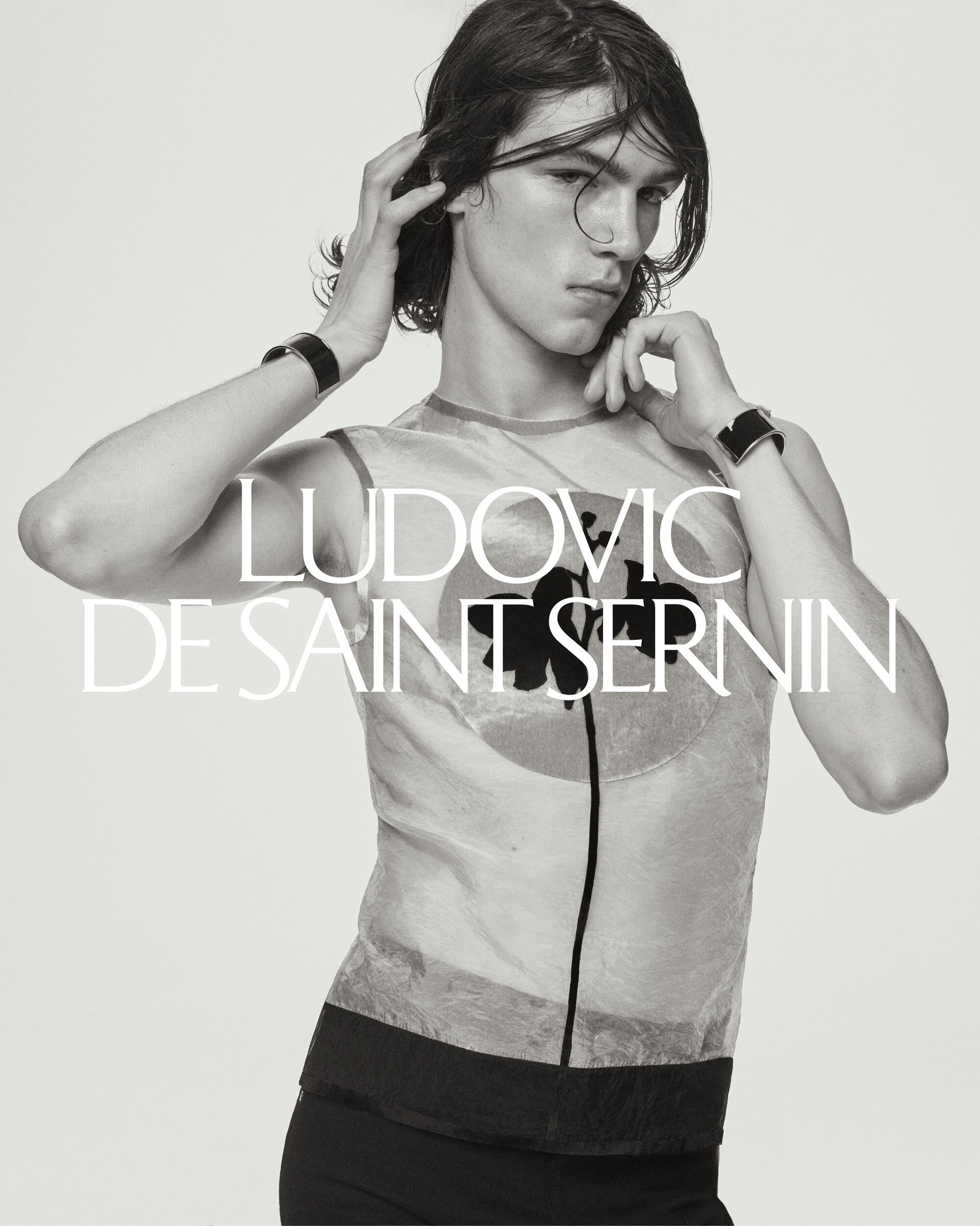 Monochrome studio portrait of a slender adult male model with medium-length hair wearing a sleeveless top and a watch posing with hands up.