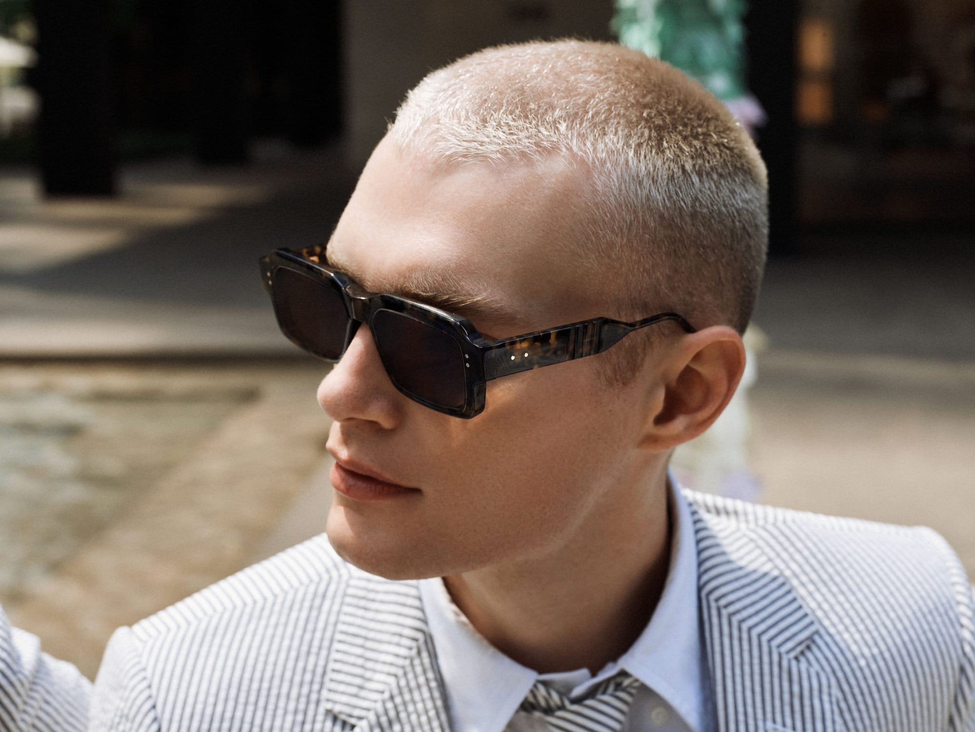 Close-up of a light-skinned man with buzzcut wearing patterned sunglasses, a white shirt and pinstripe blazer in natural light.
