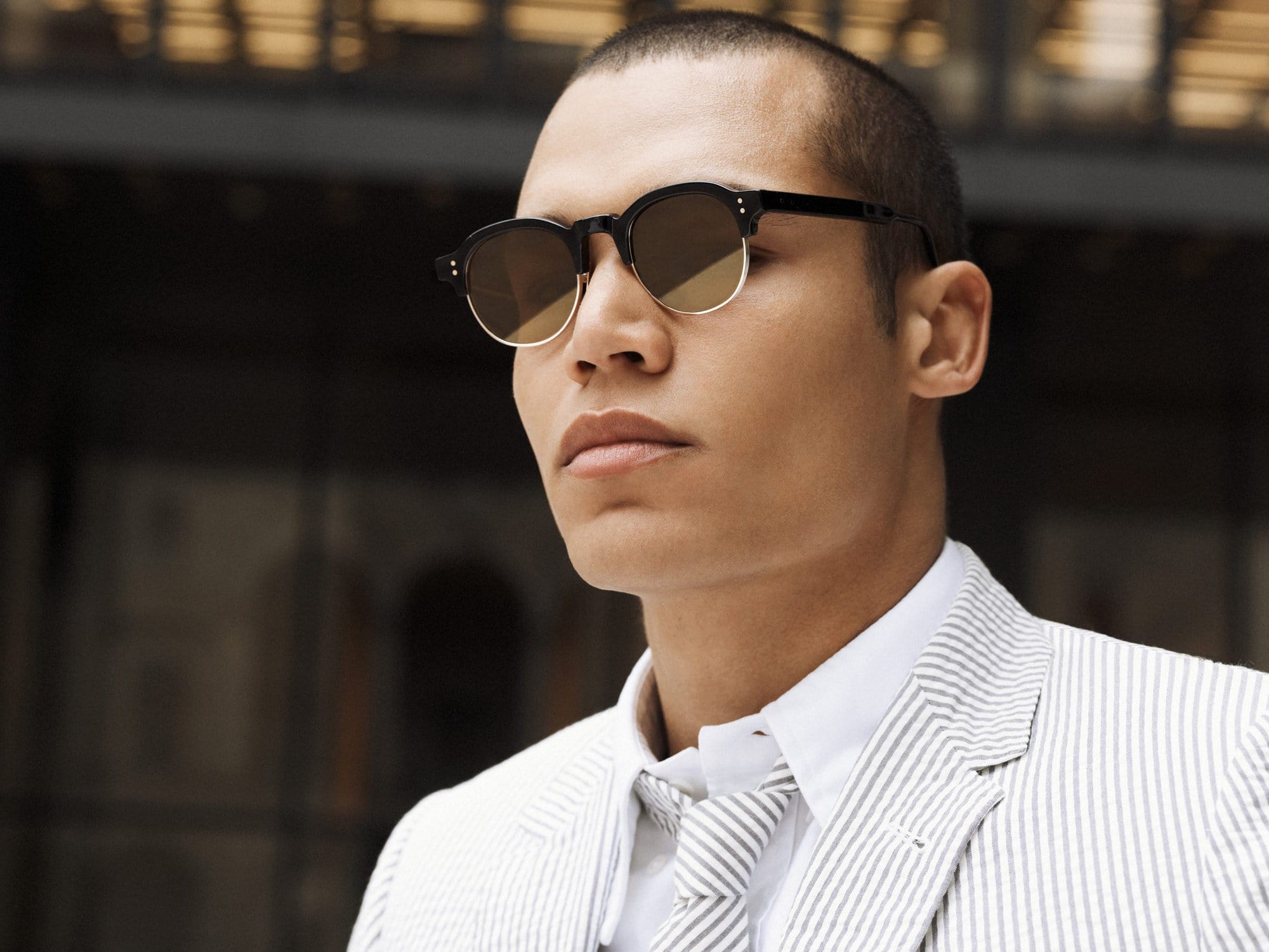 Portrait of an adult male model wearing round sunglasses, a white collared shirt, and a grey and white striped suit and tie.