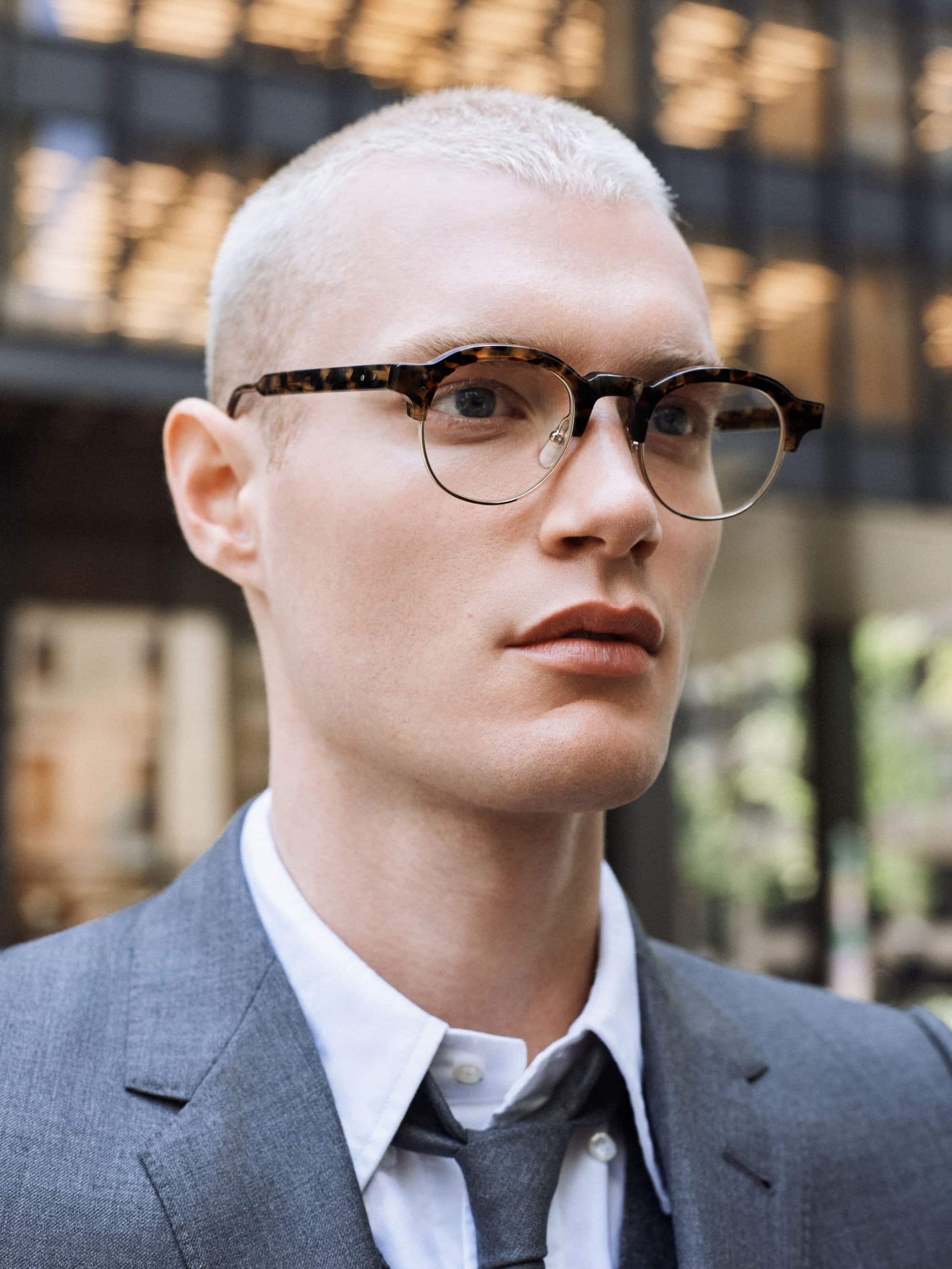 Portrait of a young adult man with buzzcut hairstyle wearing tortoiseshell glasses and a grey suit against a blurred outdoor background.