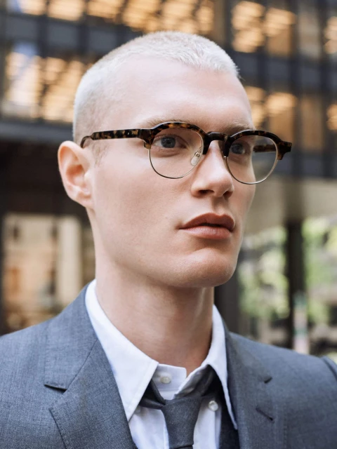 Portrait of a young adult man with buzzcut hairstyle wearing tortoiseshell glasses and a grey suit against a blurred outdoor background.
