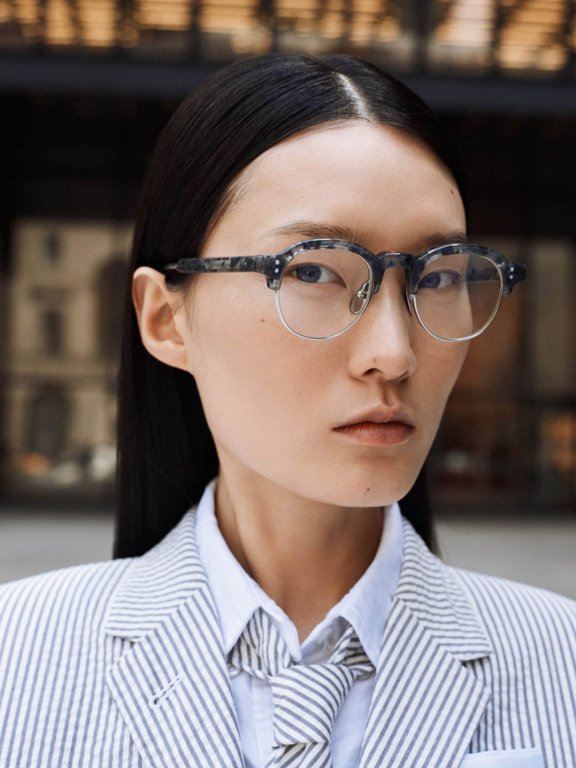 Close-up of an Asian woman with dark hair wearing tortoise shell glasses, a striped suit, tie and white shirt in an outdoor setting.