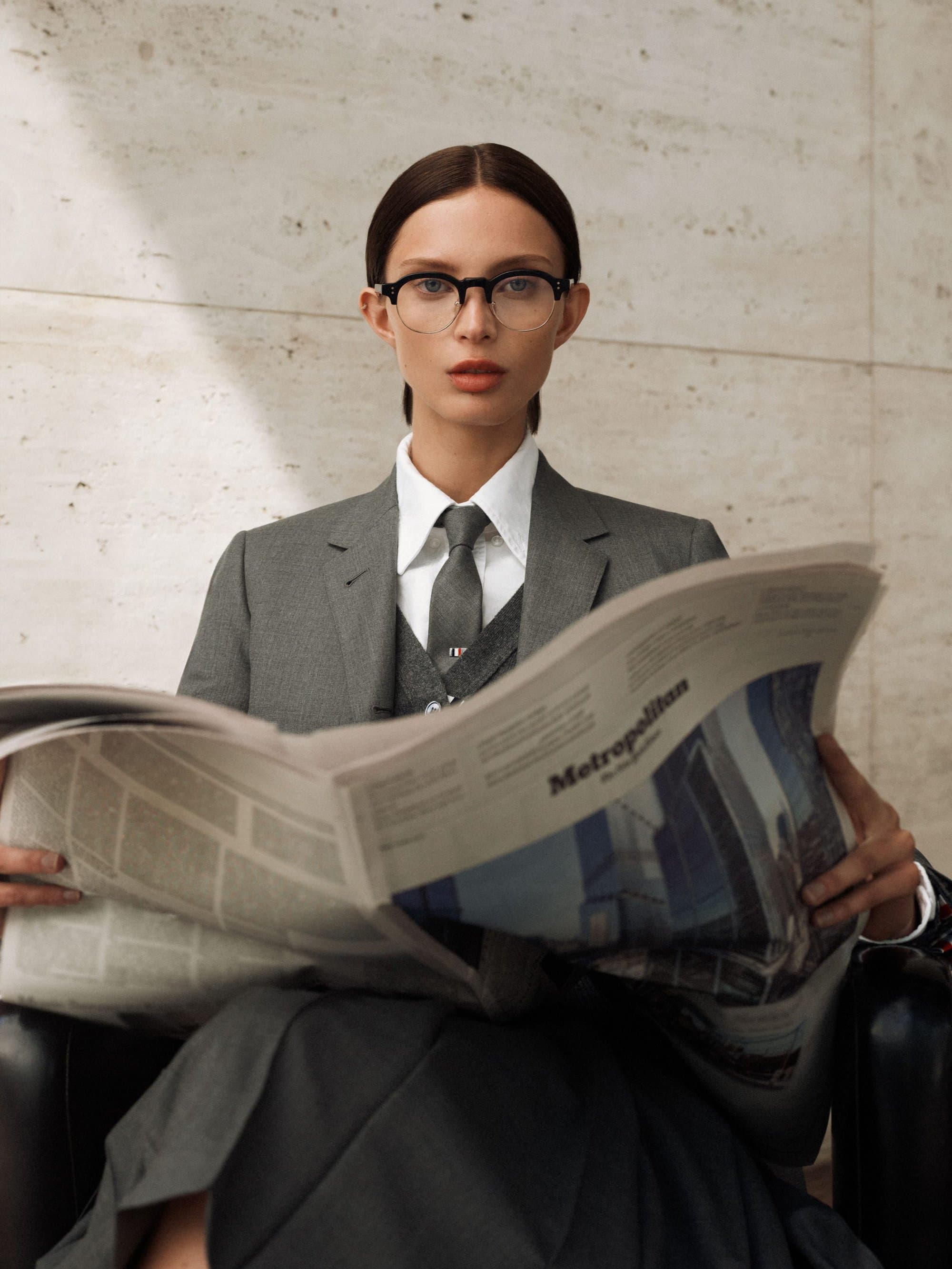 Stylish woman in a grey suit and glasses sits reading a newspaper in a bright interior with a neutral-colored stone wall.