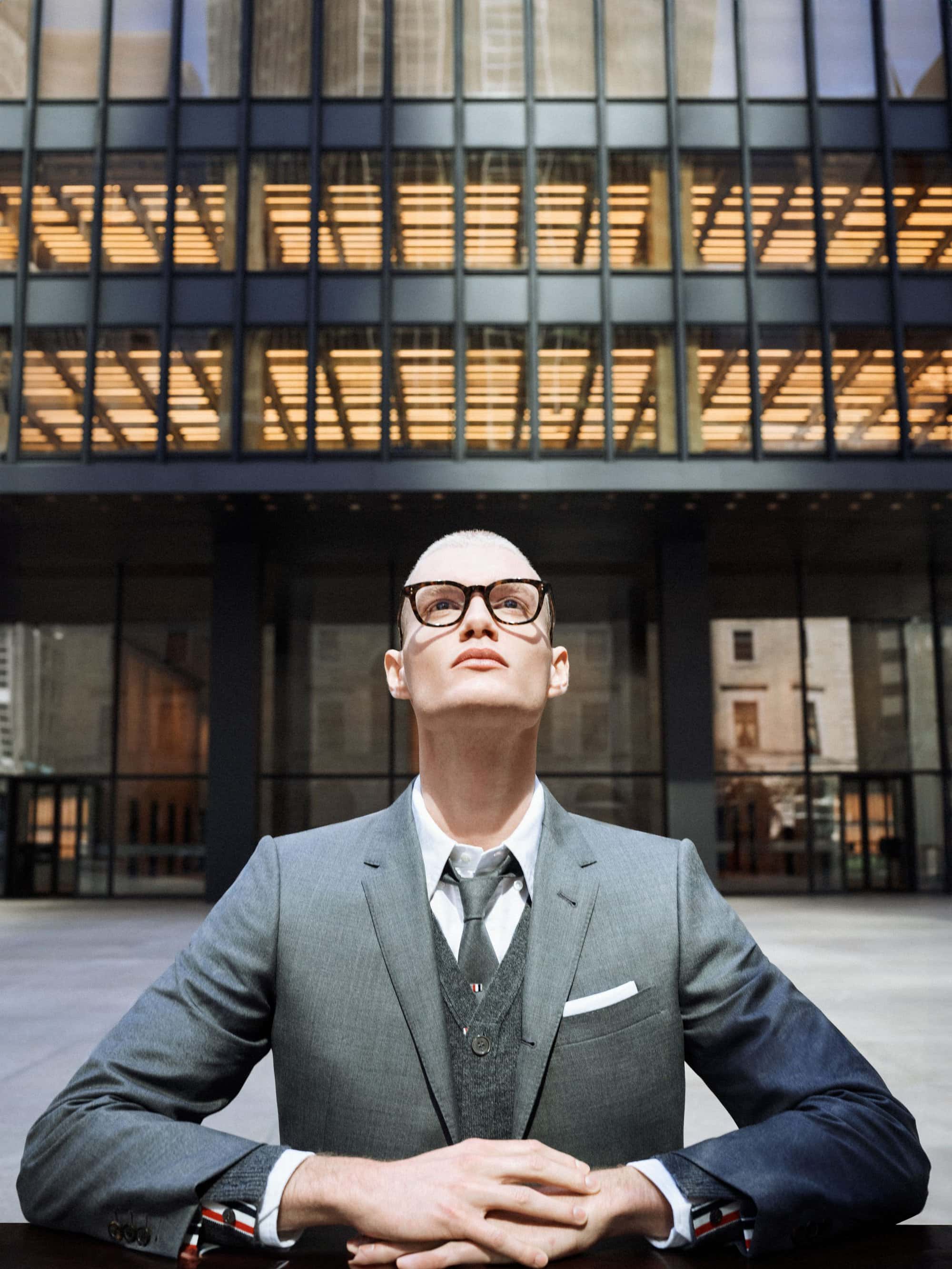 Adult male in grey suit and glasses stands outside of modern building with grid-like windows, looking upwards with neutral expression.