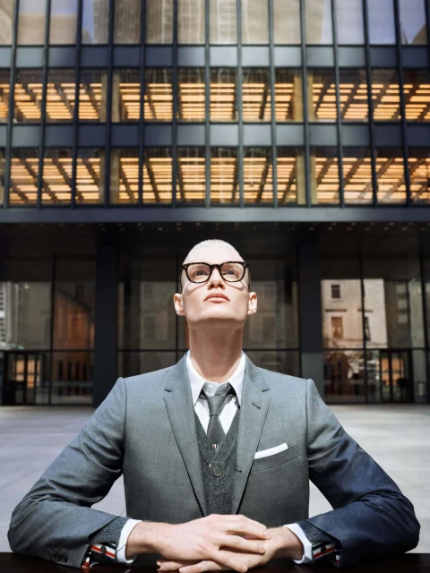 Adult male in grey suit and glasses stands outside of modern building with grid-like windows, looking upwards with neutral expression.
