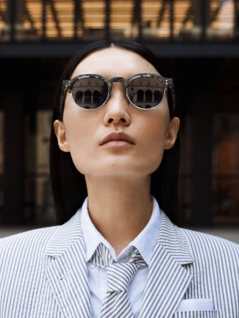 Woman in striped blazer and sunglasses stands against a dark, multi-windowed building with reflections of architecture visible in lenses.