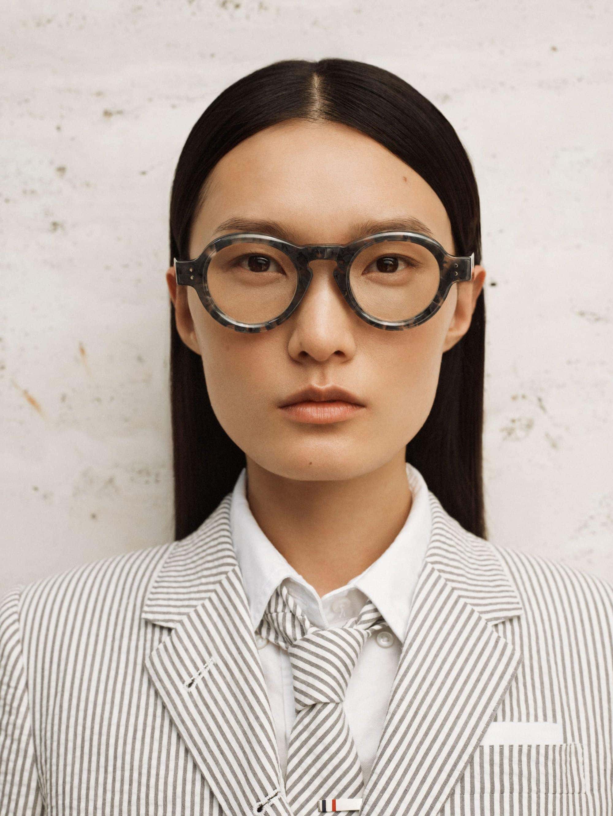 Portrait of a woman wearing eyeglasses and a seersucker suit, gazing directly at the camera against a textured background in a fashion setting.