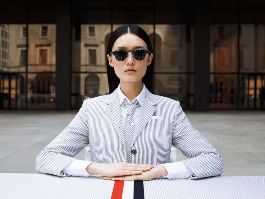 An Asian woman with long hair poses outdoors wearing sunglasses, a striped blazer, a shirt, and a tie, seated at a striped table.