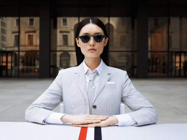 An Asian woman with long hair poses outdoors wearing sunglasses, a striped blazer, a shirt, and a tie, seated at a striped table.