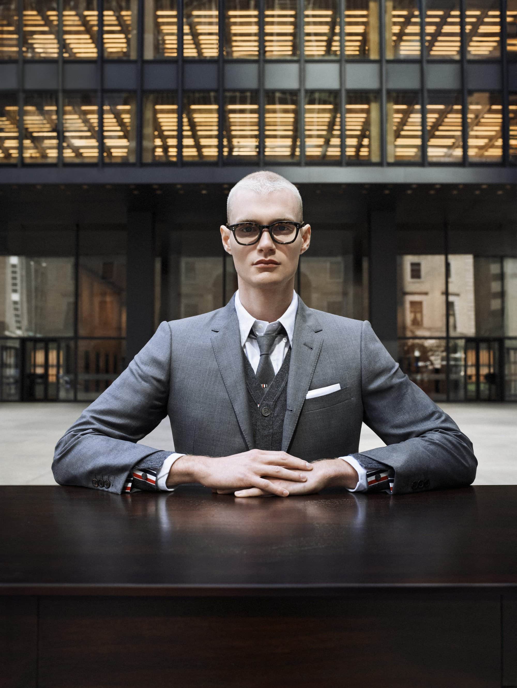 Man in gray suit and glasses sitting at a table with a modern building facade in the background, presenting a formal and architectural scene.