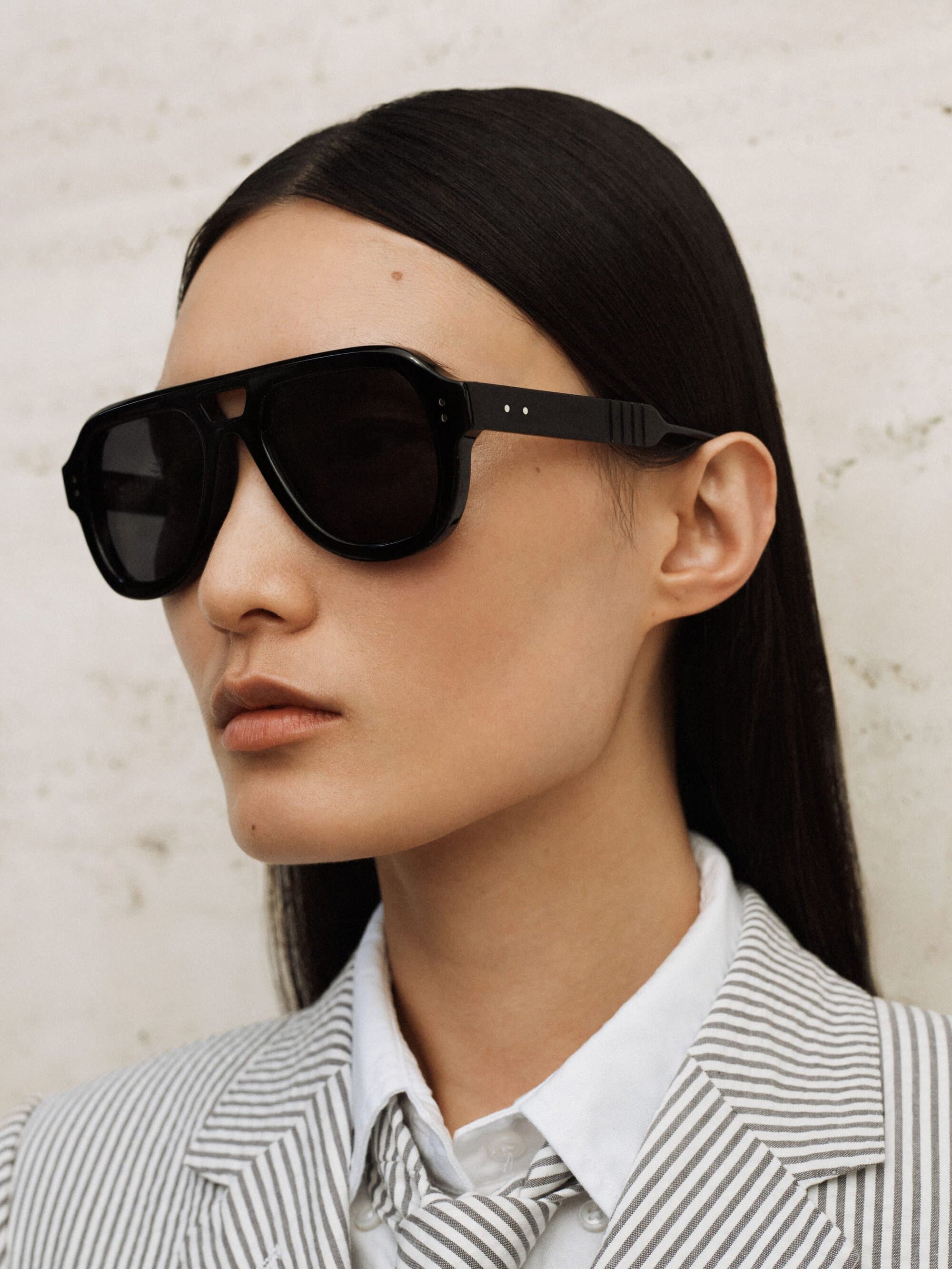 Close-up of a person with long dark hair wearing black aviator sunglasses and a white and grey pinstripe blazer with a matching necktie.