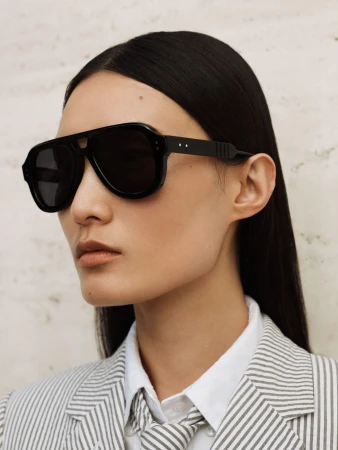 Close-up of a person with long dark hair wearing black aviator sunglasses and a white and grey pinstripe blazer with a matching necktie.