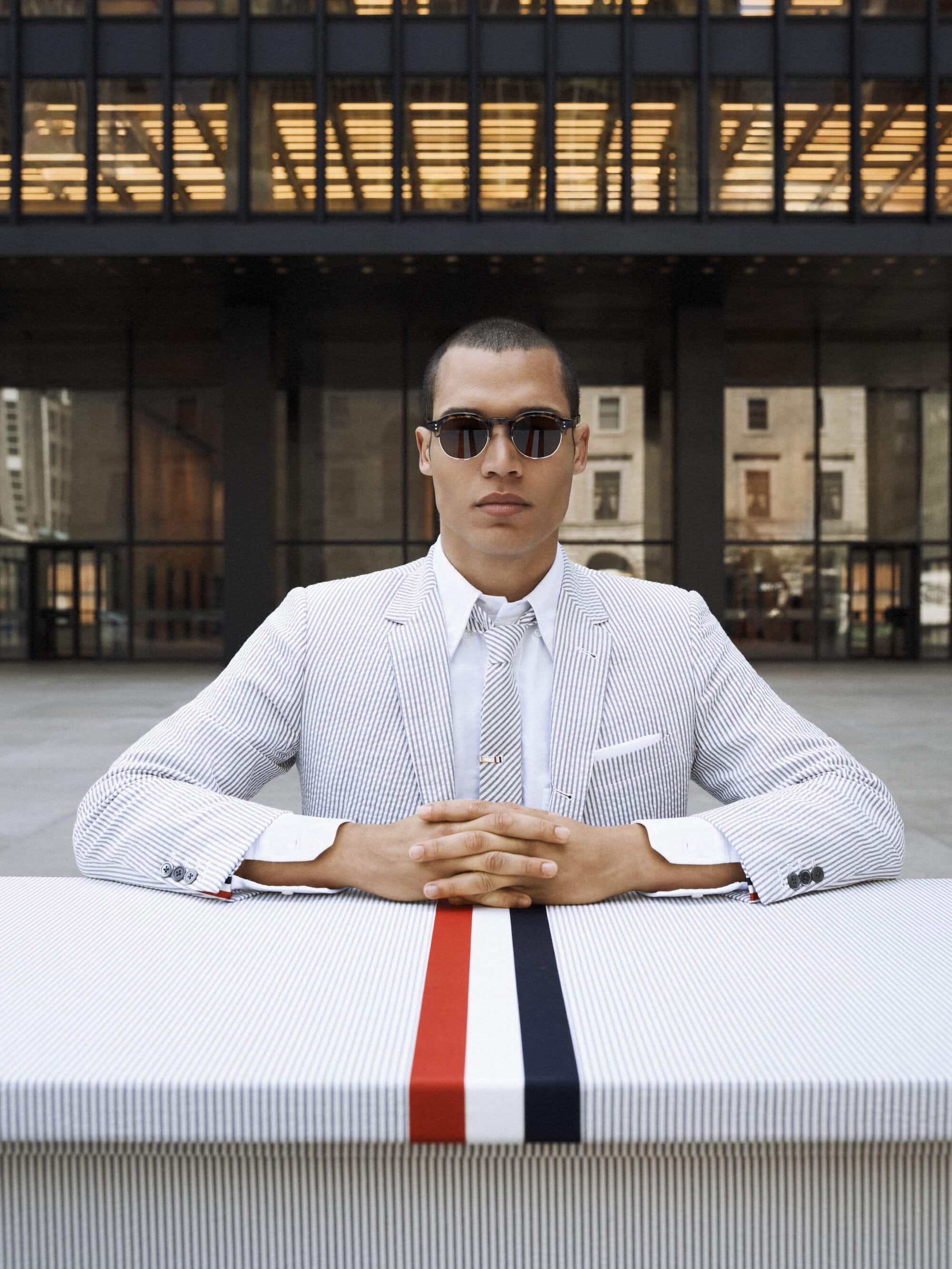 A man in a pinstripe suit and sunglasses poses behind a striped table in front of a modern glass building in an urban setting.