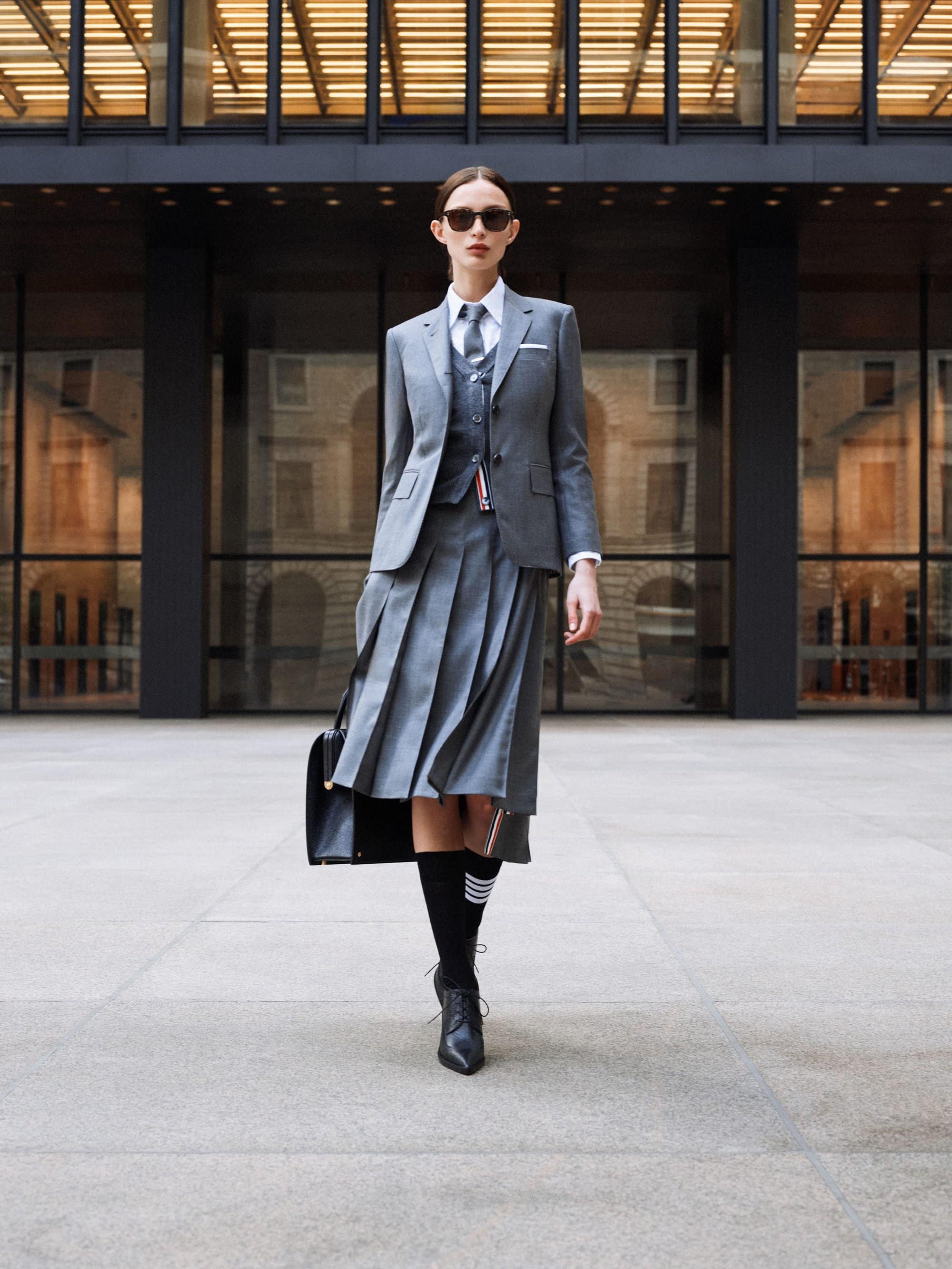 Woman in a gray suit with pleated skirt and sunglasses stands in front of a modern building, holding a black purse and wearing black boots.
