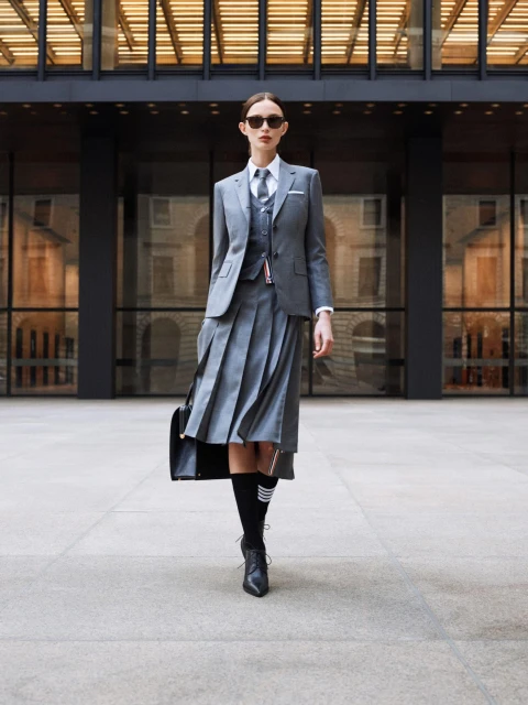 Woman in a gray suit with pleated skirt and sunglasses stands in front of a modern building, holding a black purse and wearing black boots.