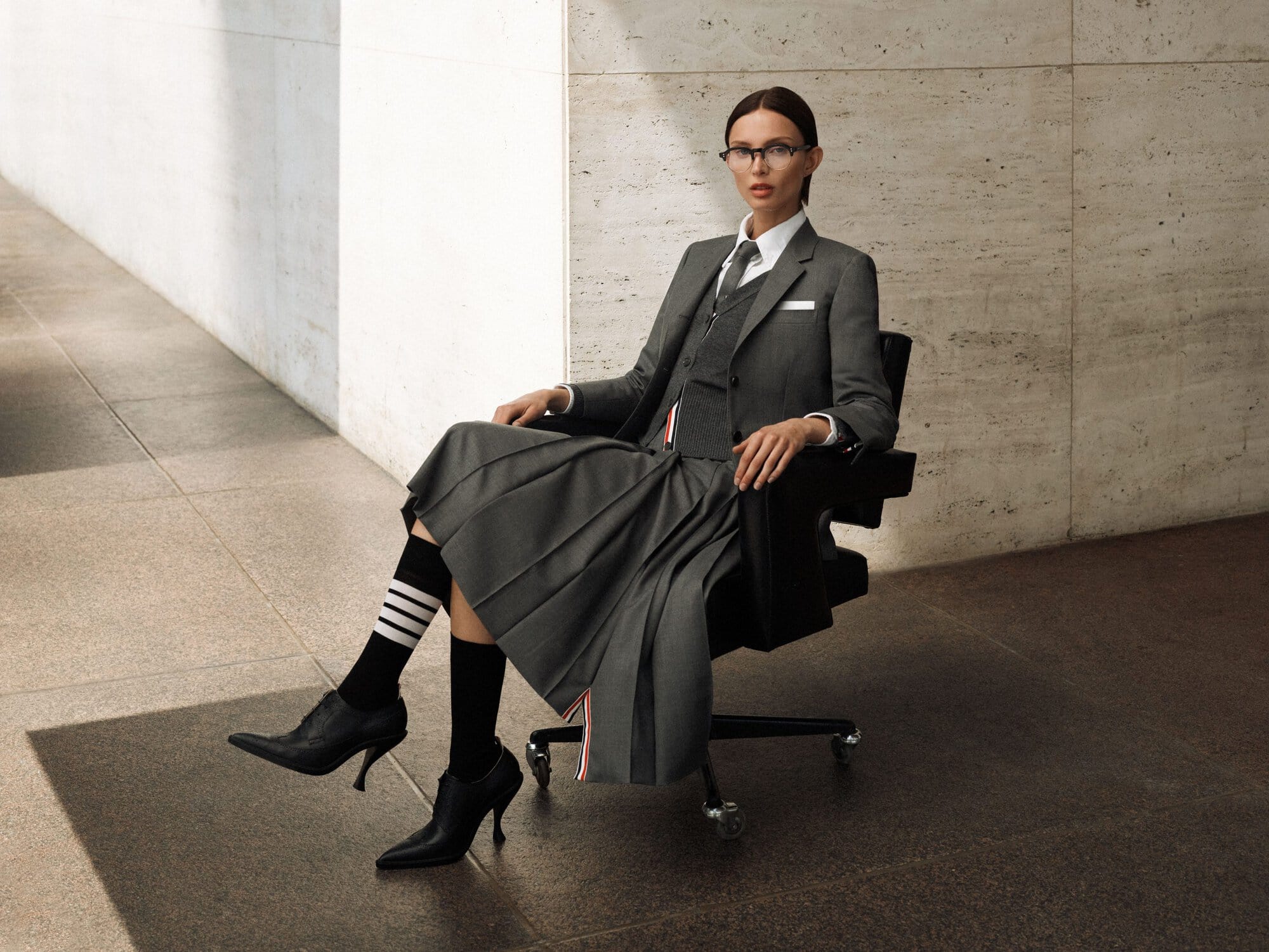 Elegant woman in a grey skirt suit sitting on a black chair in front of a marble wall. She wears black heels and striped socks.