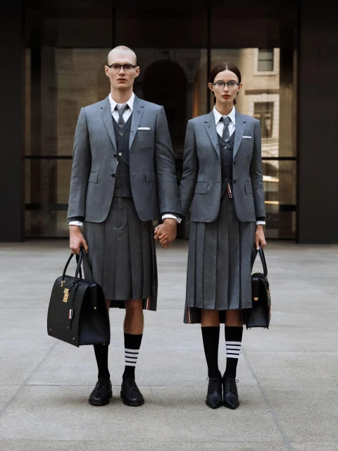 Stylish couple in grey suits and pleated skirts holding hands outdoors, each carrying a black bag, with a modern building backdrop.