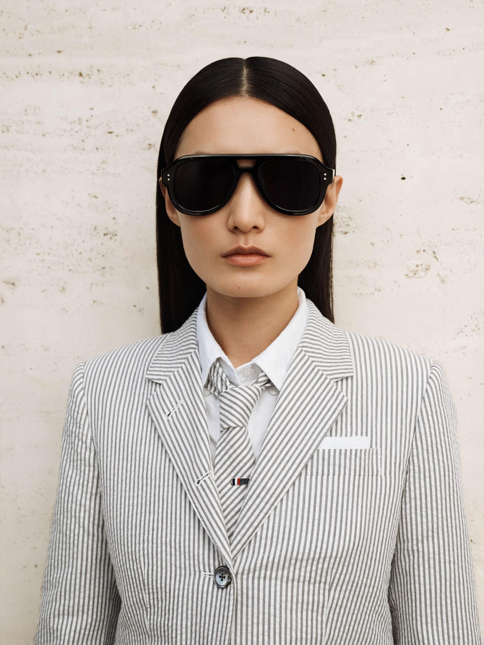 Stylish woman in a pinstripe suit and black sunglasses gazes at the camera against a textured neutral backdrop, exuding sophisticated fashion.