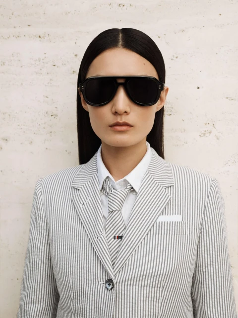 Stylish woman in a pinstripe suit and black sunglasses gazes at the camera against a textured neutral backdrop, exuding sophisticated fashion.