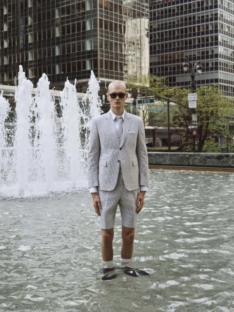 A man in a gray seersucker suit and sunglasses stands in a fountain in a city with tall buildings in the background, posing for a fashion shoot.