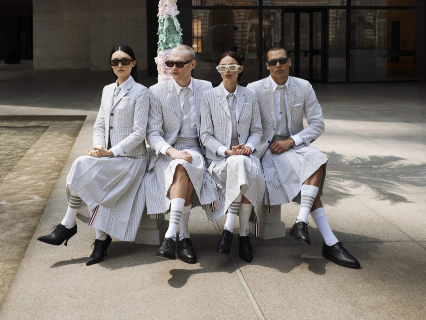 Group of four diverse adult models seated in suits and skirts with socks, sunglasses, and dress shoes in an outdoor urban setting.