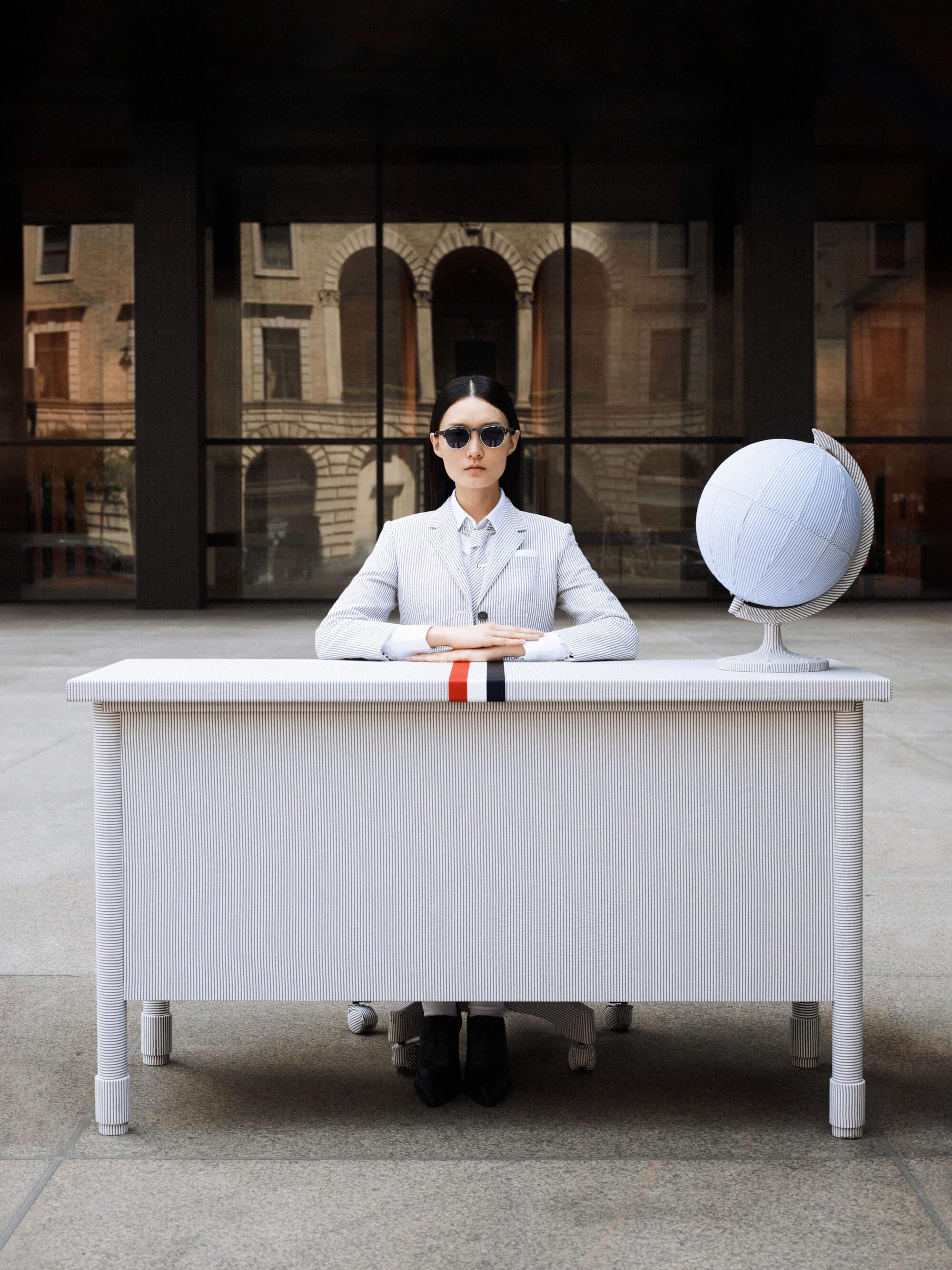 Fashion shot of a woman in a light grey suit sitting at a white desk with a globe, sunglasses, against the backdrop of a building.