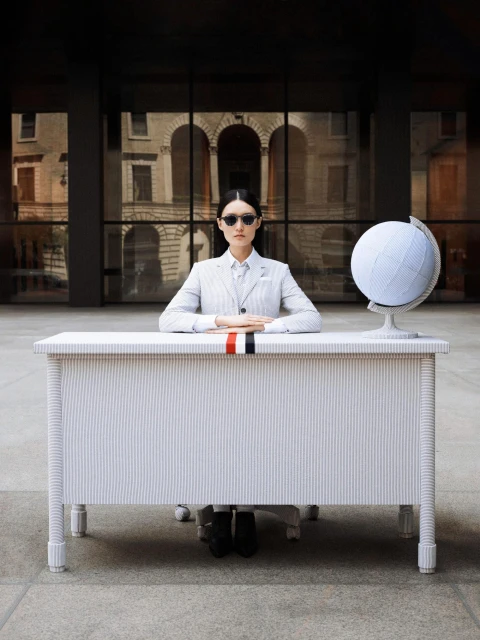 Fashion shot of a woman in a light grey suit sitting at a white desk with a globe, sunglasses, against the backdrop of a building.