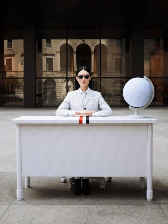 Fashion shot of a woman in a light grey suit sitting at a white desk with a globe, sunglasses, against the backdrop of a building.
