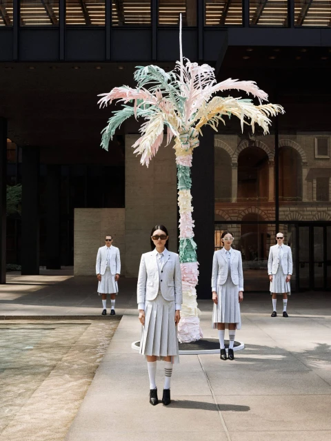 Four fashion models in light-grey skirt suits stand in front of a pastel-colored palm tree sculpture outside a modern building.
