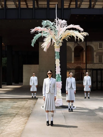 Four fashion models in light-grey skirt suits stand in front of a pastel-colored palm tree sculpture outside a modern building.